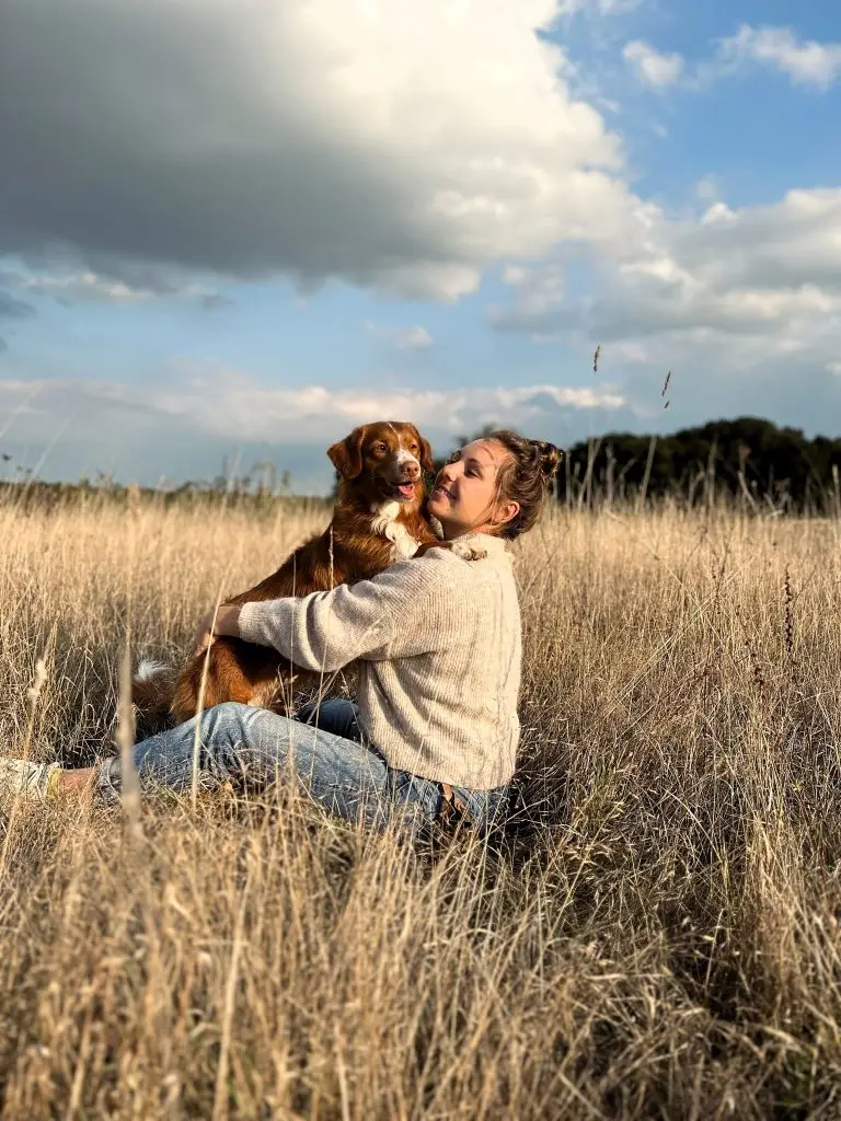 Jeune femme assise dans un champ d'herbes hautes serrant tendrement son chien Nova Scotia Duck Tolling Retriever contre elle.